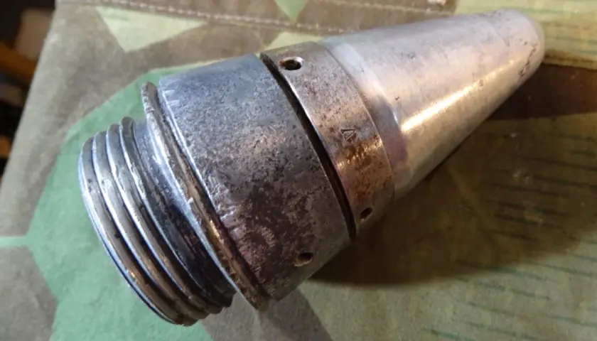 Ein metallisches Teil, das an eine **Flakgranate** erinnert, mit Gewinde und spitz zulaufendem Ende, liegt auf Tarnstoff.