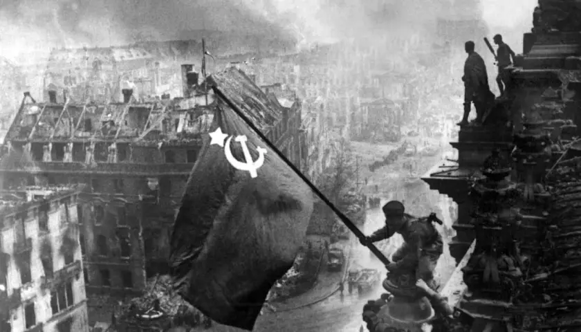 Sowjetische Soldaten hissen die rote Fahne über dem zerstörten Reichstag 1945. Rauch und Trümmer prägen das Bild Berlins.