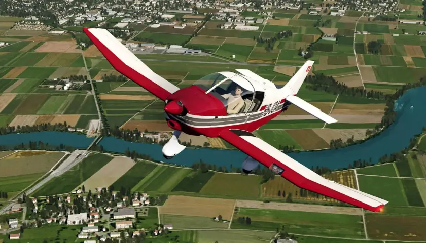 Ein roter und weißer Robin DR 400 fliegt über grüne Felder und einen Fluss.