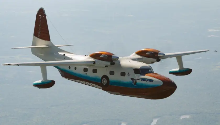 Grumman Mallard Wasserflugzeug fliegt über bewaldetem Gebiet. Das Flugzeug hat eine weiße und braune Lackierung mit blauen Akzenten.