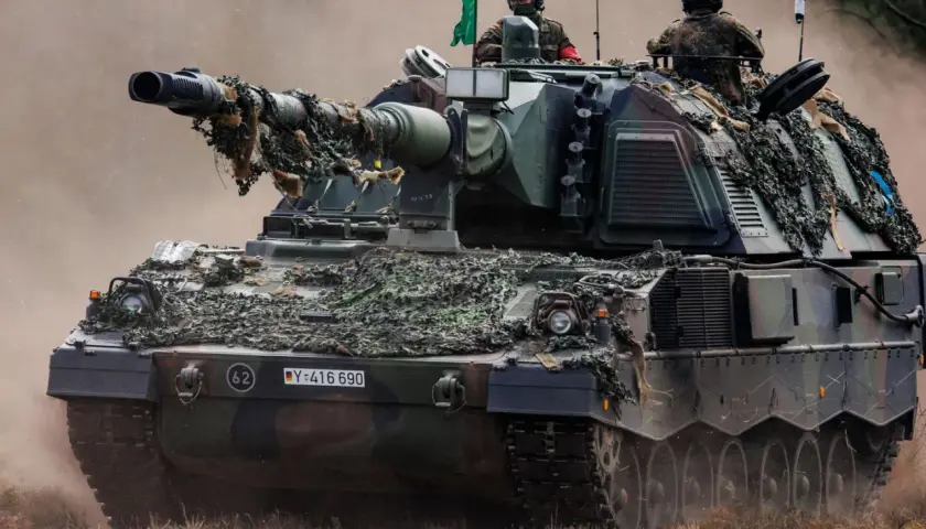 Eine Panzerhaubitze 3000 mit Tarnnetz fährt durch staubiges Gelände. Zwei Soldaten sind auf dem Turm zu sehen.