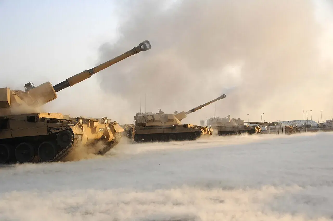 Mehrere Panzerhaubitzen, wie die 90, feuern in einer staubigen, rauchigen Umgebung.
