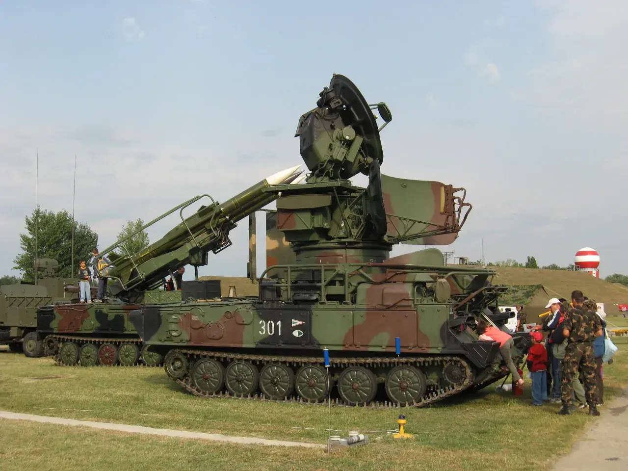 Ein Raketenwerfer SA-6 mit Raketen und Radar steht auf einem Feld. Kinder und Erwachsene betrachten die militärische Ausrüstung.