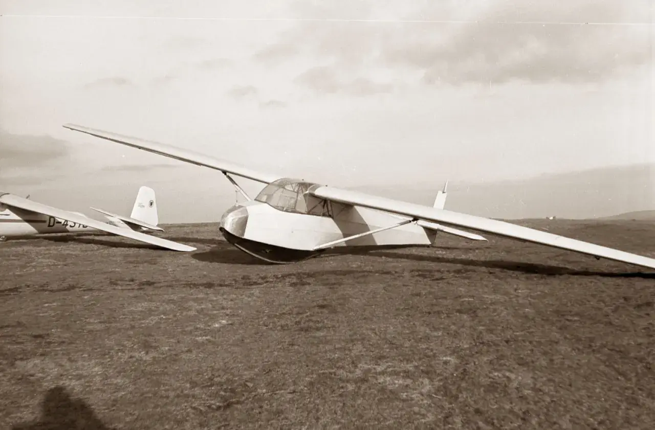 Ein Segelflugzeug vom Typ Musger MG 19 steht auf einer Wiese. Ein weiteres Flugzeug ist im Hintergrund zu sehen.