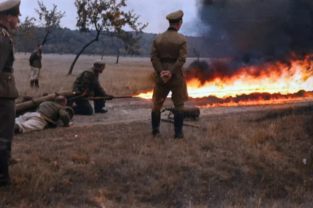 Soldaten im 2. Weltkrieg üben den Einsatz eines Flammenwerfers. Ein Mann zielt mit dem Gerät, während ein anderer auf dem Boden liegt.