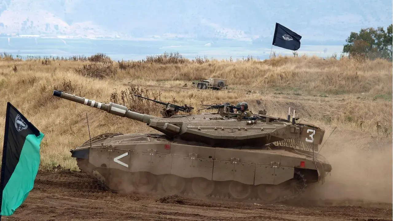 Ein Merkava Panzer fährt durch staubiges Gelände, begleitet von zwei Fahnen.