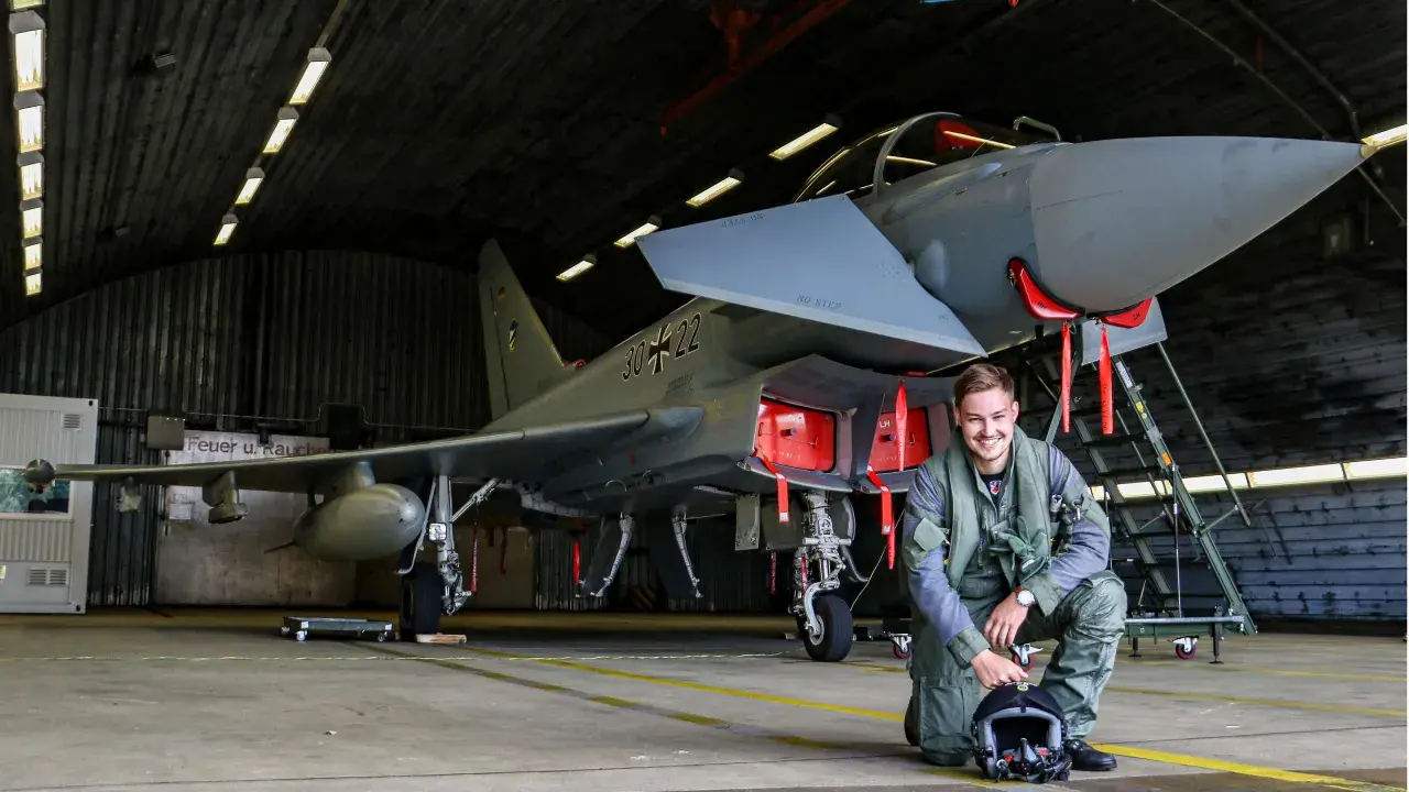 Ein junger F16 Pilot in Uniform kniet neben seinem Jet in einer Flugzeughalle.