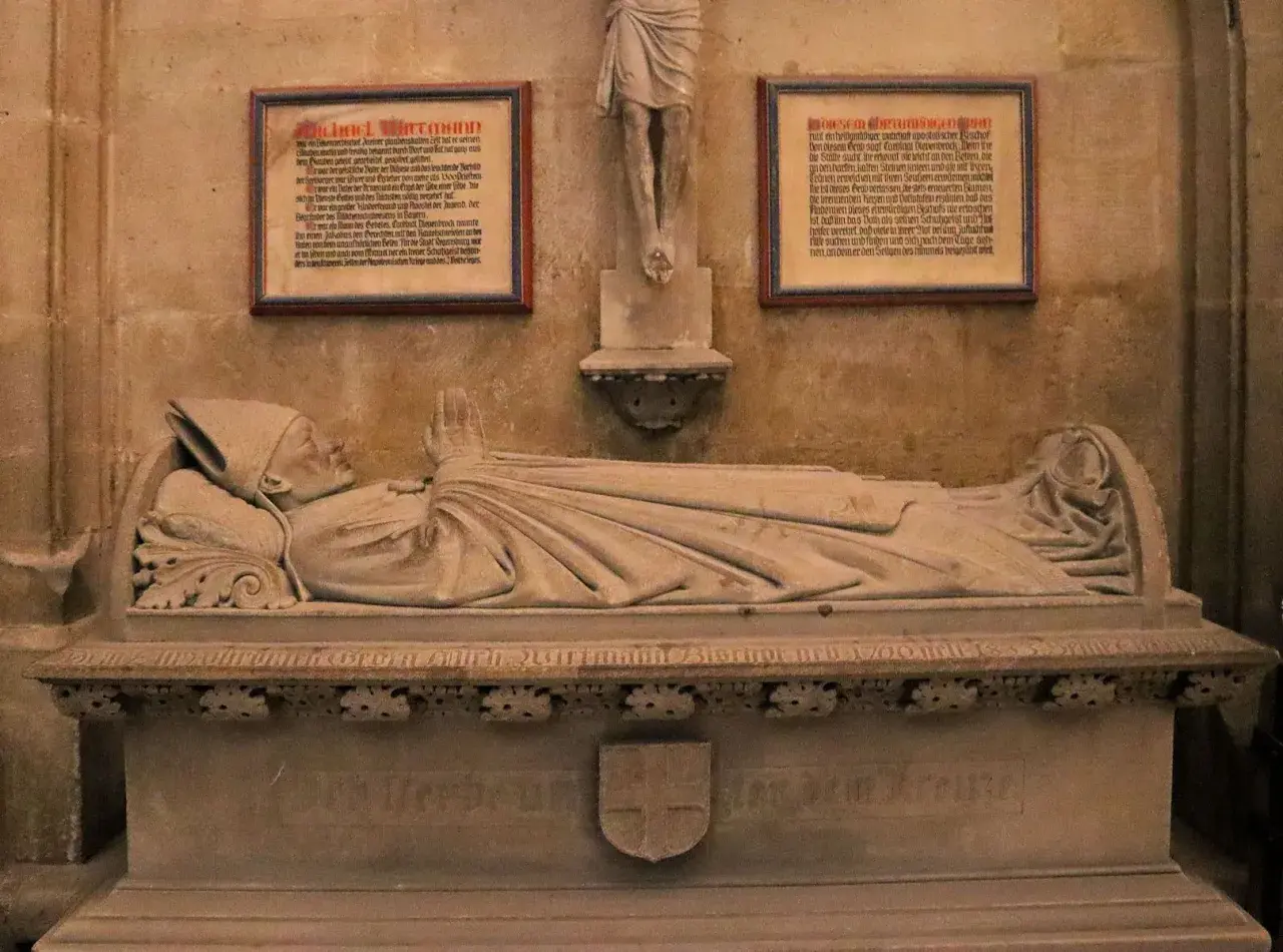 Das Michael Wittmann Grab zeigt eine liegende Steinfigur eines Bischofs mit gefalteten Händen. Tafeln mit Texten flankieren die Skulptur.