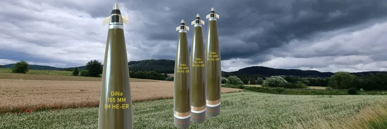 Vier Geschosse des Kalibers 155 mm stehen im Vordergrund vor einer ländlichen Kulisse mit Feldern und bewaldeten Hügeln unter dramatischem Himmel.