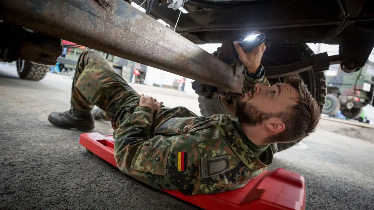 Ein Soldat, ein Unteroffizier ohne Portepee, prüft mit einer Lampe das Fahrgestell eines Fahrzeugs.