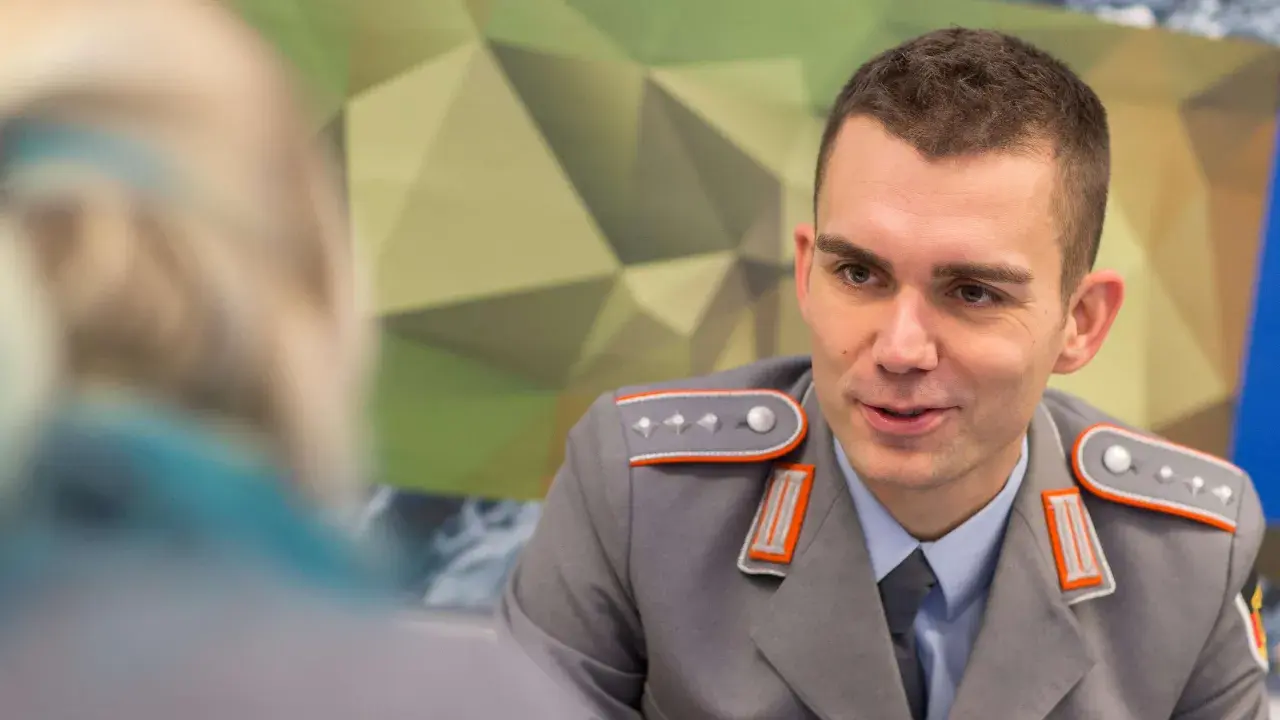 Ein junger Fachoffizier der Bundeswehr im Gespräch, trägt eine graue Uniform mit orangefarbenen Akzenten und Schulterklappen.