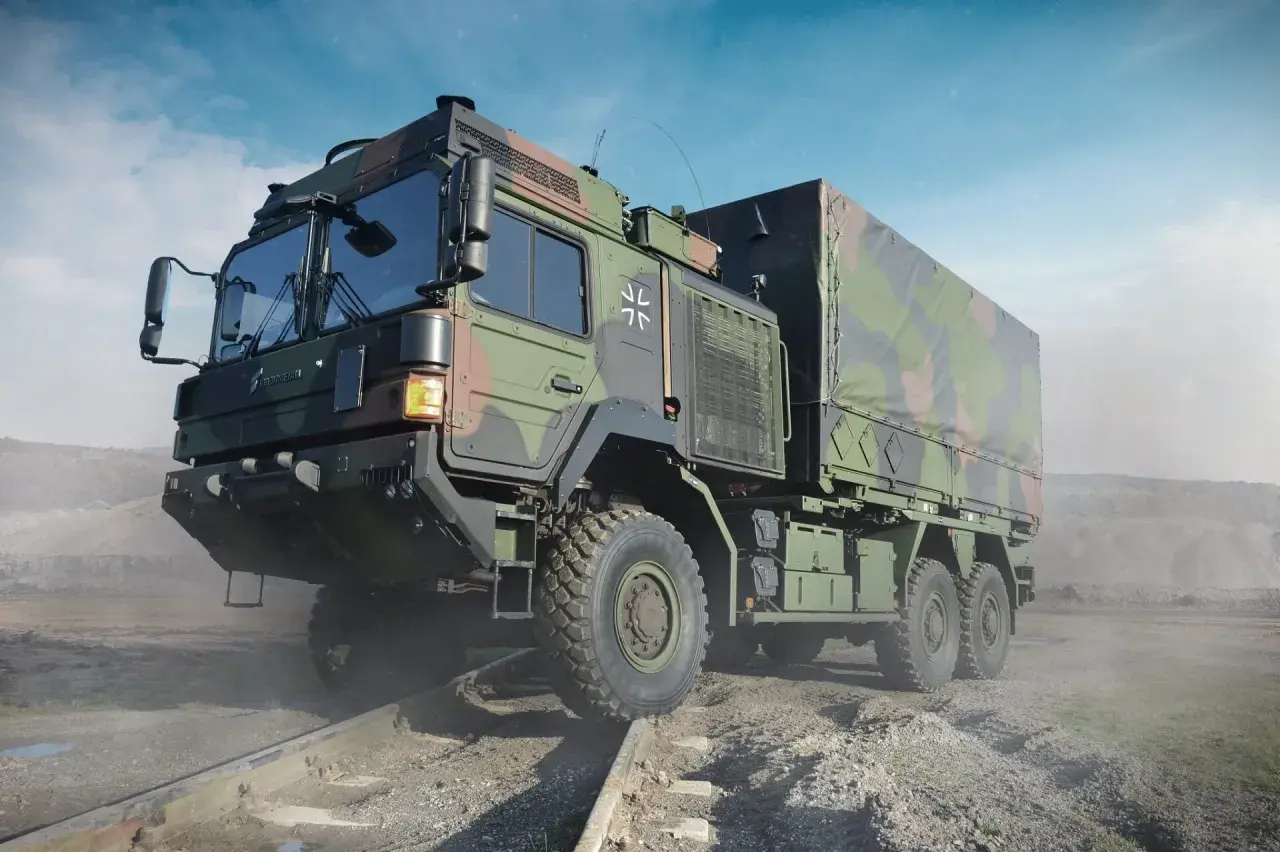 Ein gepanzerter LKW der Bundeswehr mit Tarnmuster fährt über Schienen in einer staubigen Landschaft.