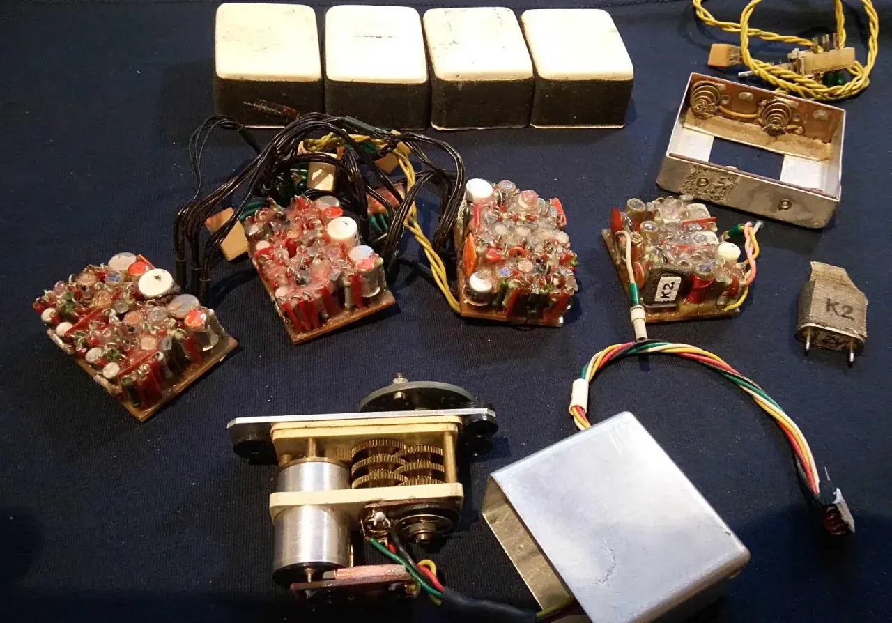 Elektronikbauteile, darunter vier weiße Kästen, mehrere Leiterplatten mit vielen Kondensatoren und Drähten, ein Motor mit Zahnrädern und ein Metallgehäuse. Vielleicht für 27 MHz Funkgeräte.