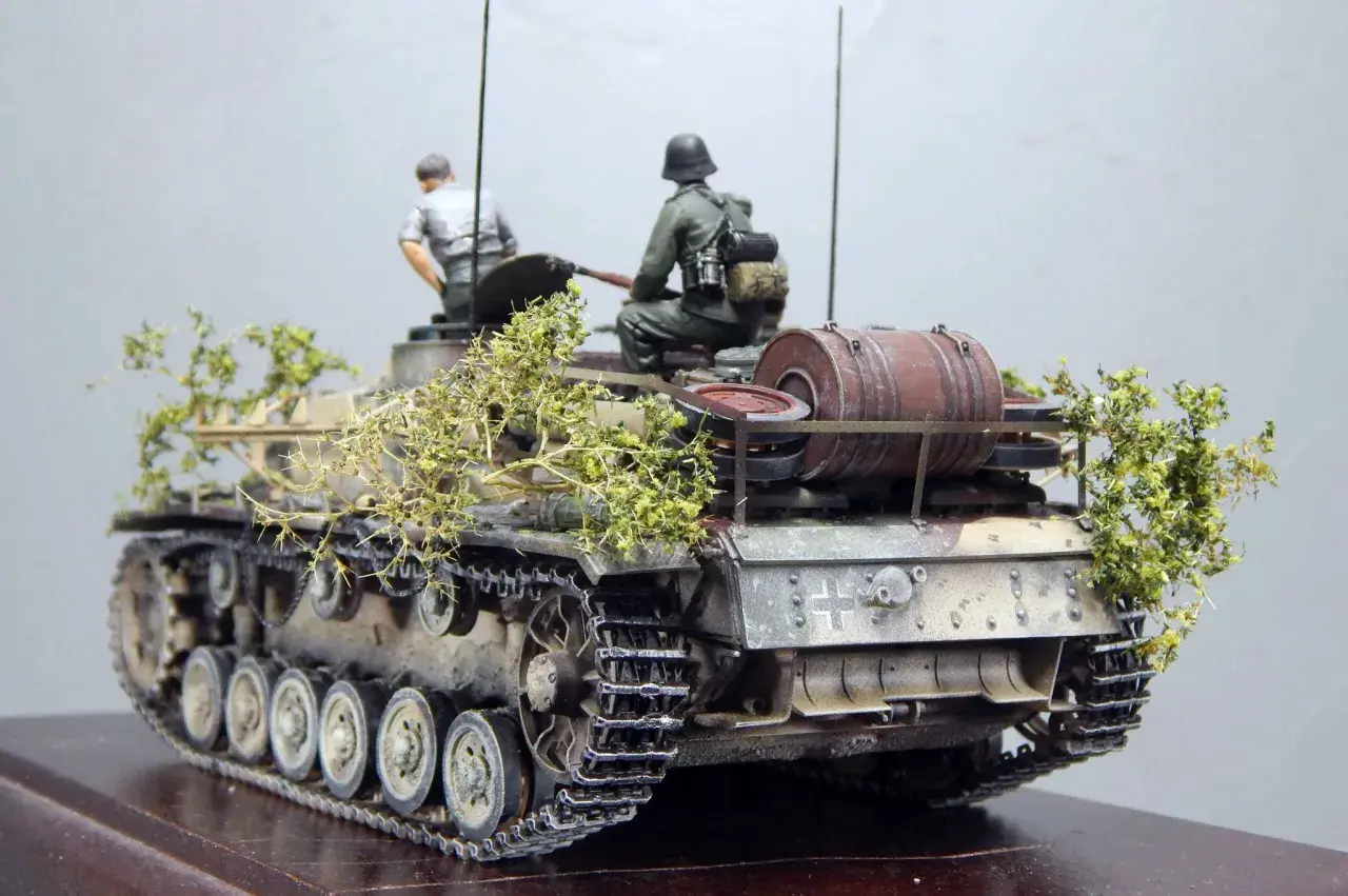 Sturmhaubitze 42 Modell mit zwei Figuren, getarnt mit Zweigen.