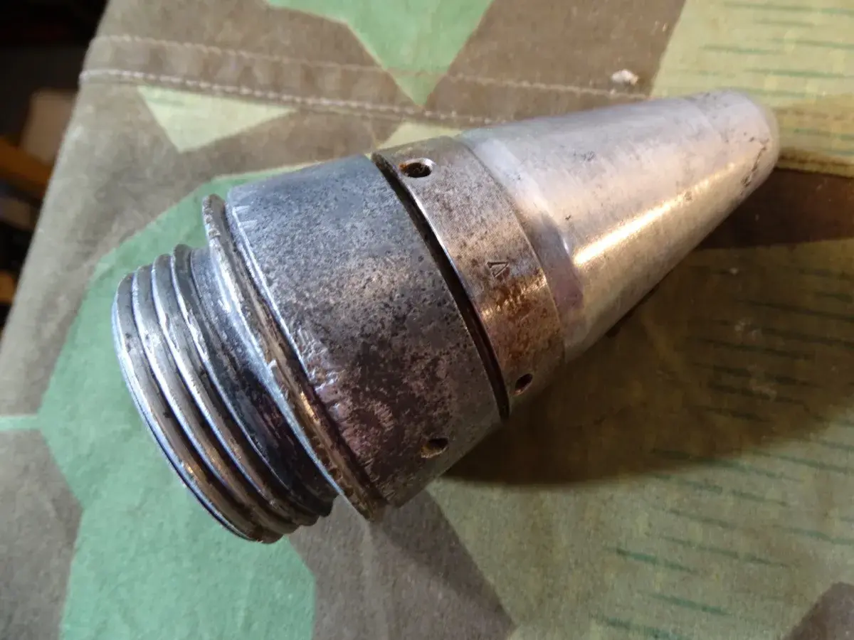 Ein metallisches Teil, das an eine **Flakgranate** erinnert, mit Gewinde und spitz zulaufendem Ende, liegt auf Tarnstoff.