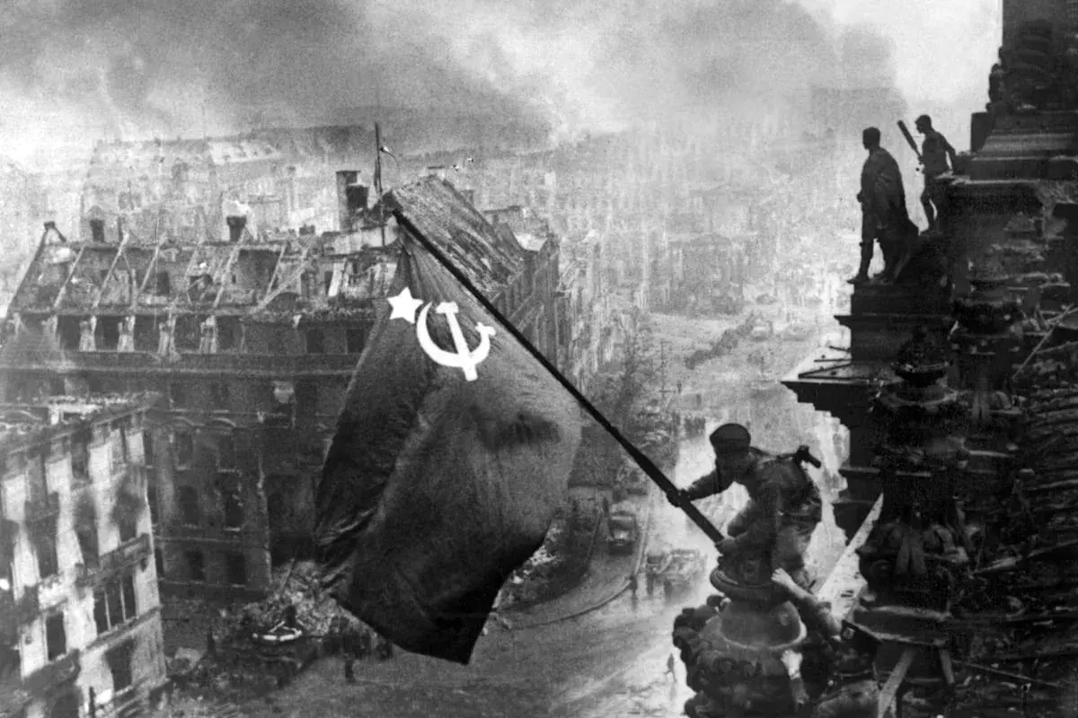 Sowjetische Soldaten hissen die rote Fahne über dem zerstörten Reichstag 1945. Rauch und Trümmer prägen das Bild Berlins.