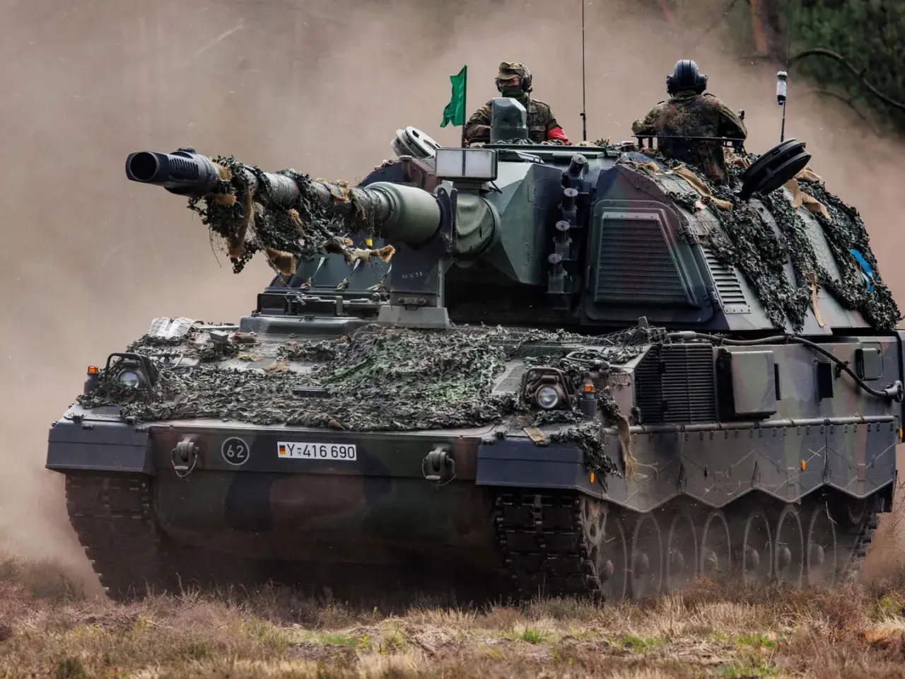 Eine Panzerhaubitze 3000 mit Tarnnetz fährt durch staubiges Gelände. Zwei Soldaten sind auf dem Turm zu sehen.