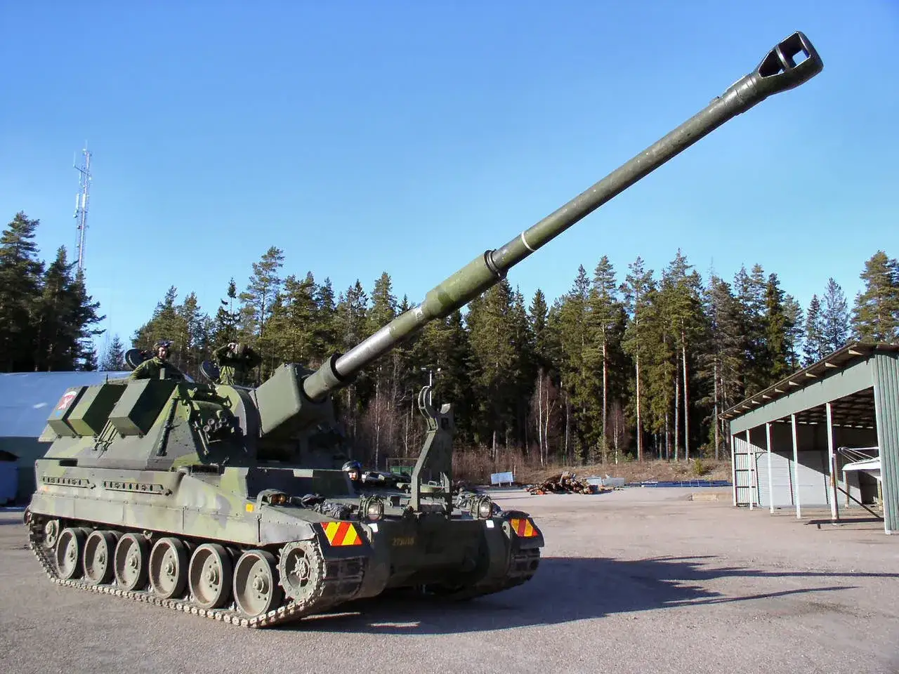 Eine 90 Haubitze steht auf einem Schotterplatz vor einem Wald. Die Kanone ist erhoben, und Soldaten sind auf dem Fahrzeug zu sehen.