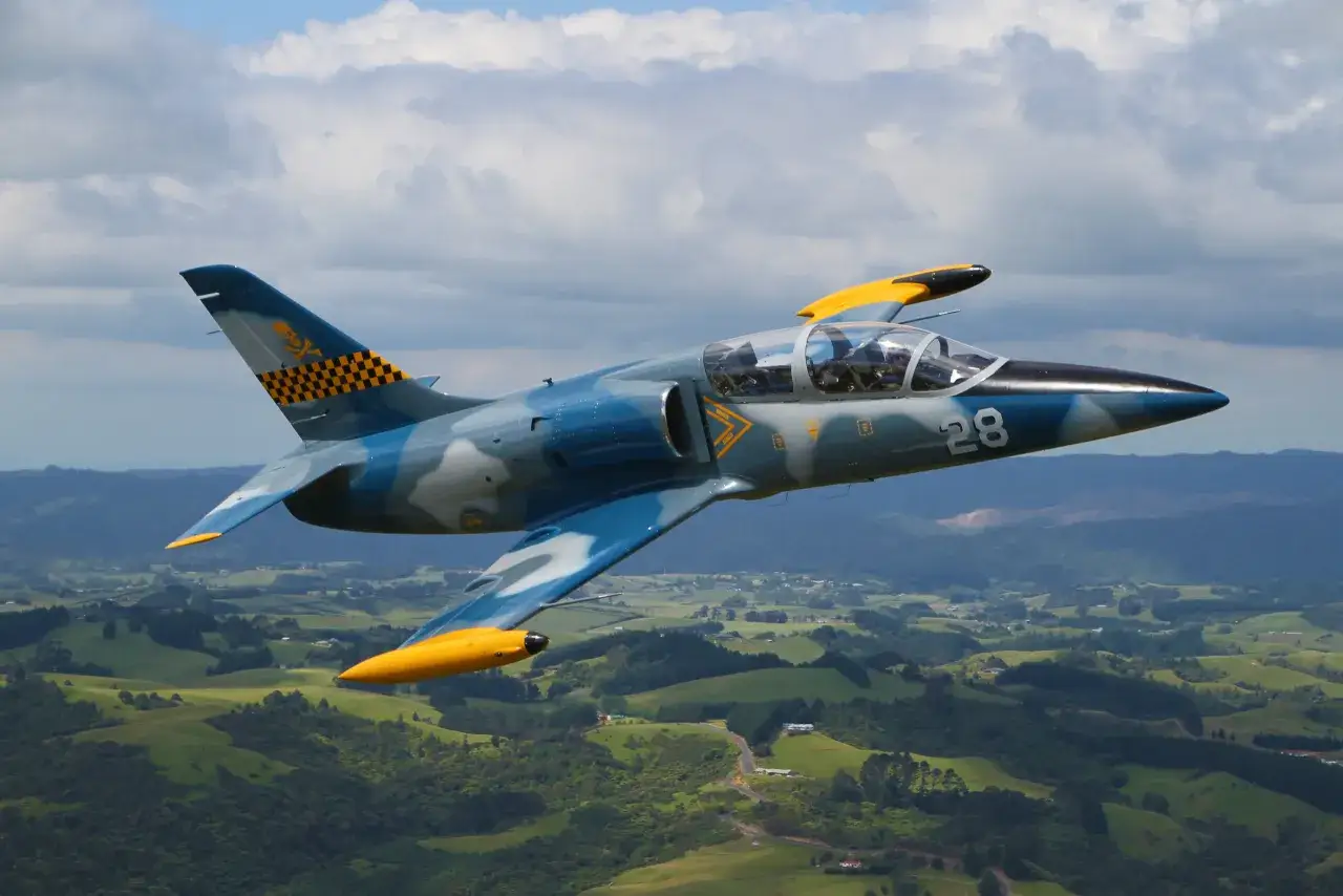 Ein L-39 Albatros Jet mit auffälliger Tarnung und gelben Akzenten fliegt über eine grüne Hügellandschaft.