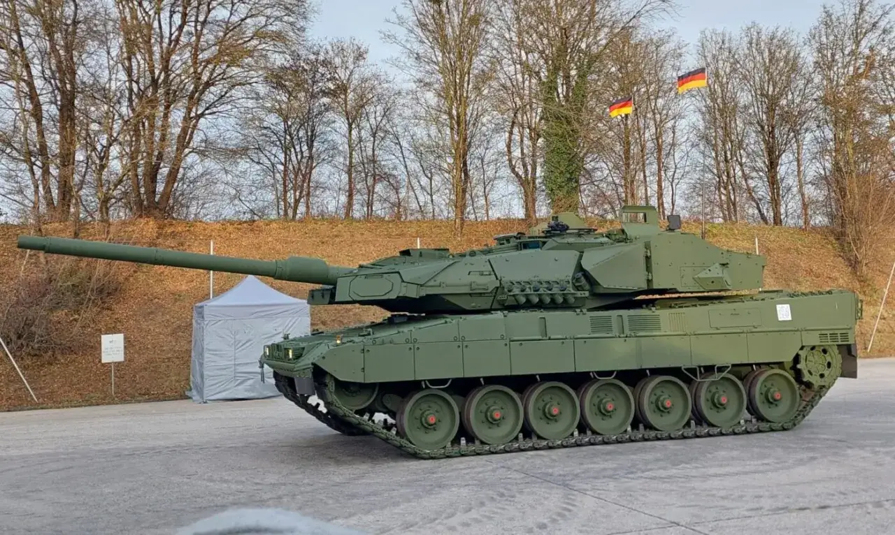 Ein grüner Leopard 2 Panzer steht im Freien. Im Hintergrund sind Bäume und zwei deutsche Flaggen zu sehen. Ein Panzer T-90 vs. Leopard 2 ist ein spannendes Duell.