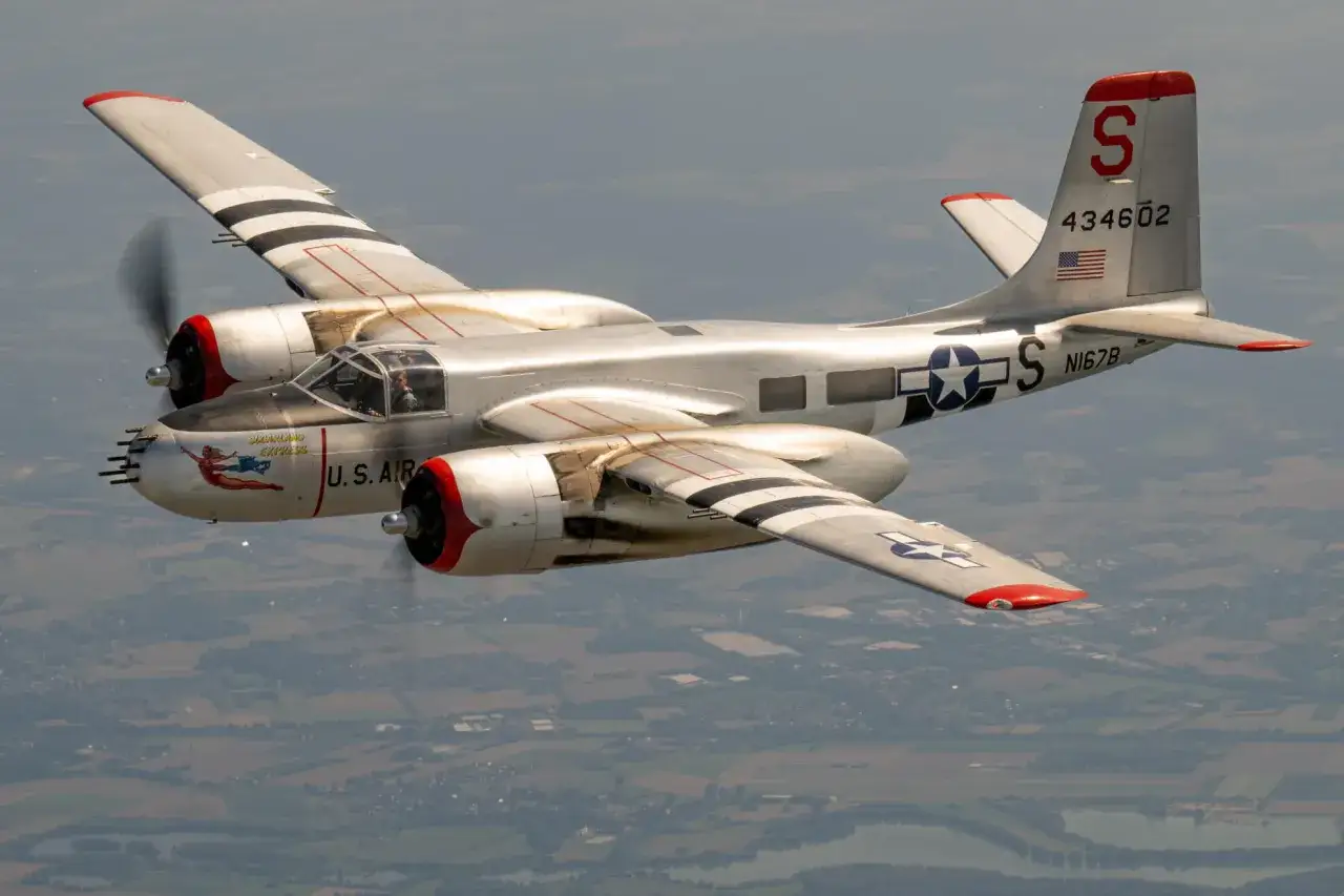 Silberne A-26 Invader mit roten Motorkapseln und schwarzen Streifen auf den Fl&uuml;geln fliegt &uuml;ber eine Landschaft.
