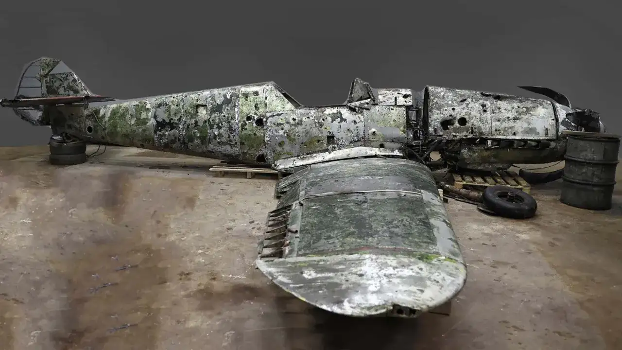 Verwitterte Bf 109 F4, stark beschädigt, mit Moosbewuchs und Einschusslöchern, liegt auf einem Holzboden.