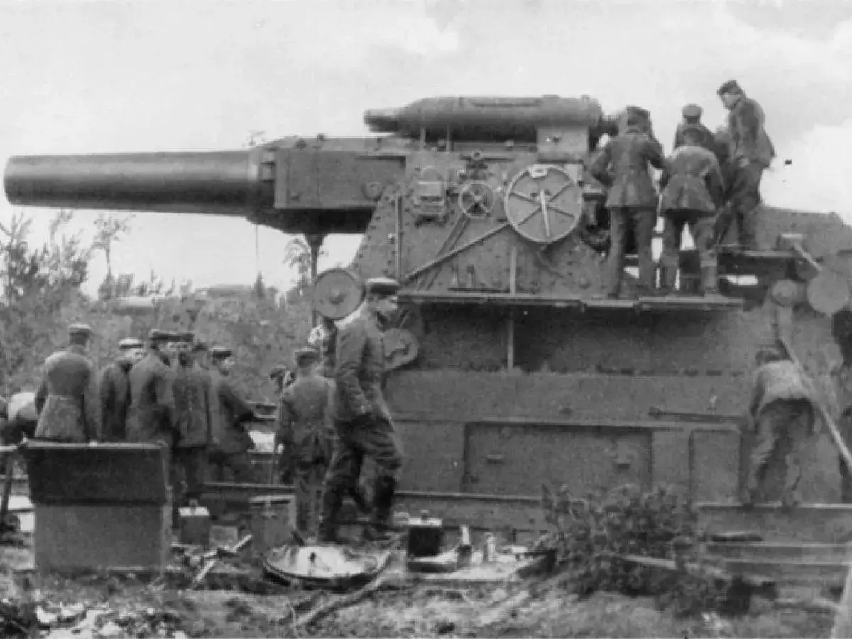 Soldaten bedienen die riesige dicke Berta Kanone. Das Geschütz ist auf einer Lafette montiert, umgeben von Vegetation und militärischem Gerät.