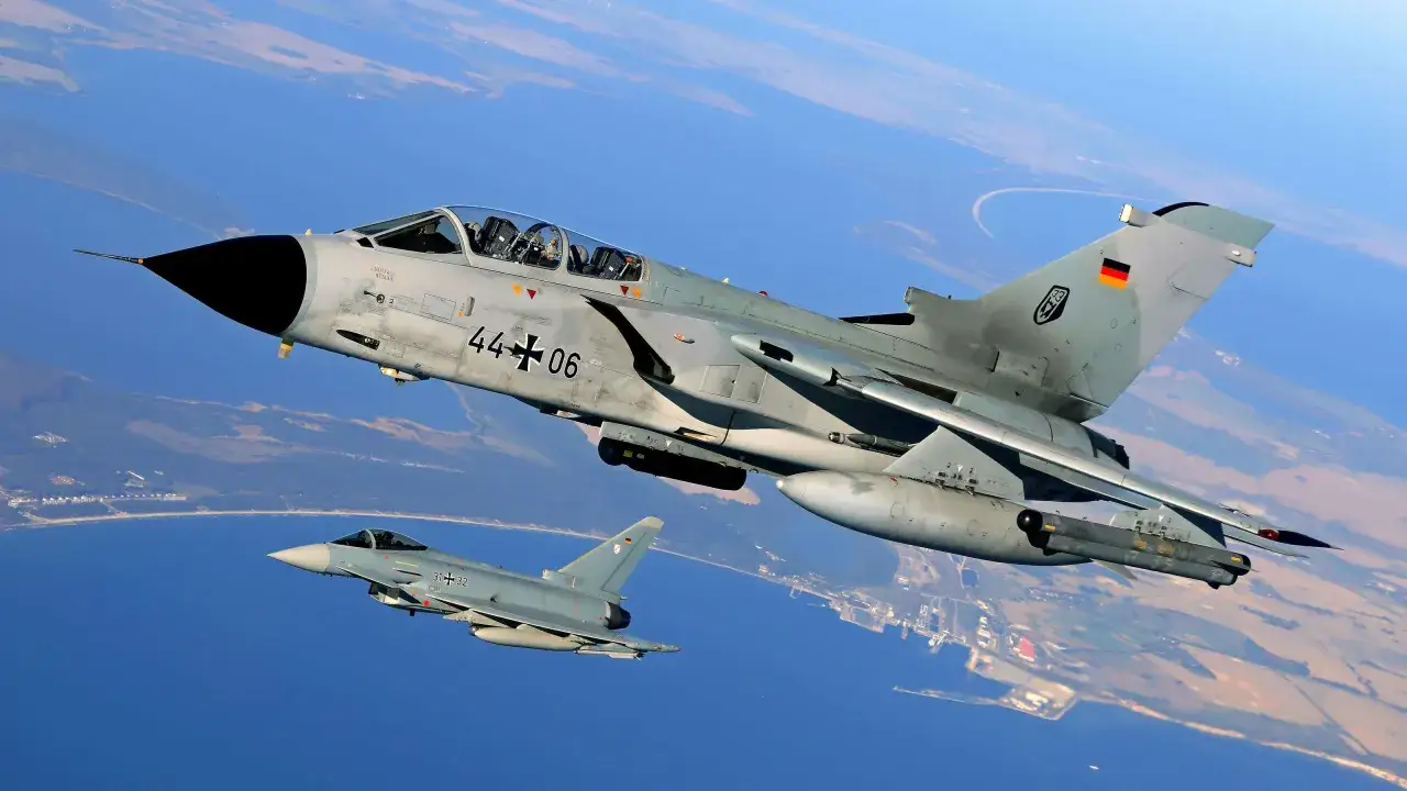 Zwei deutsche Kampfflugzeuge, ein PA-200 Tornado und ein Eurofighter, fliegen über Küstenlandschaft.