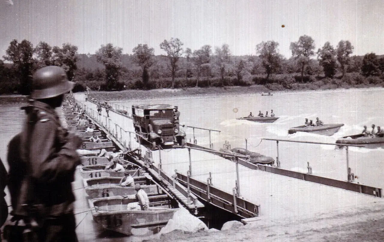 Ein deutscher Kfz-Pionier &uuml;berwacht den Aufbau einer Pontonbr&uuml;cke &uuml;ber einen Fluss. Boote mit Soldaten kreuzen auf dem Wasser.
