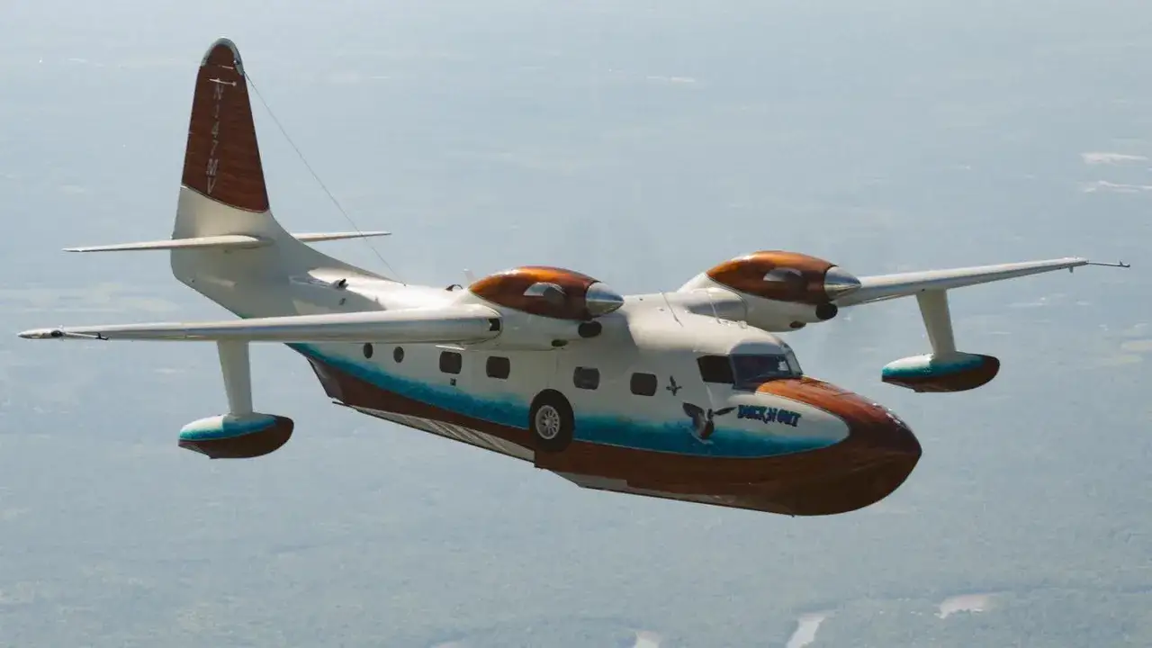 Grumman Mallard Wasserflugzeug fliegt über grünen Wäldern. Das Flugzeug hat eine markante Holzverkleidung und den Schriftzug