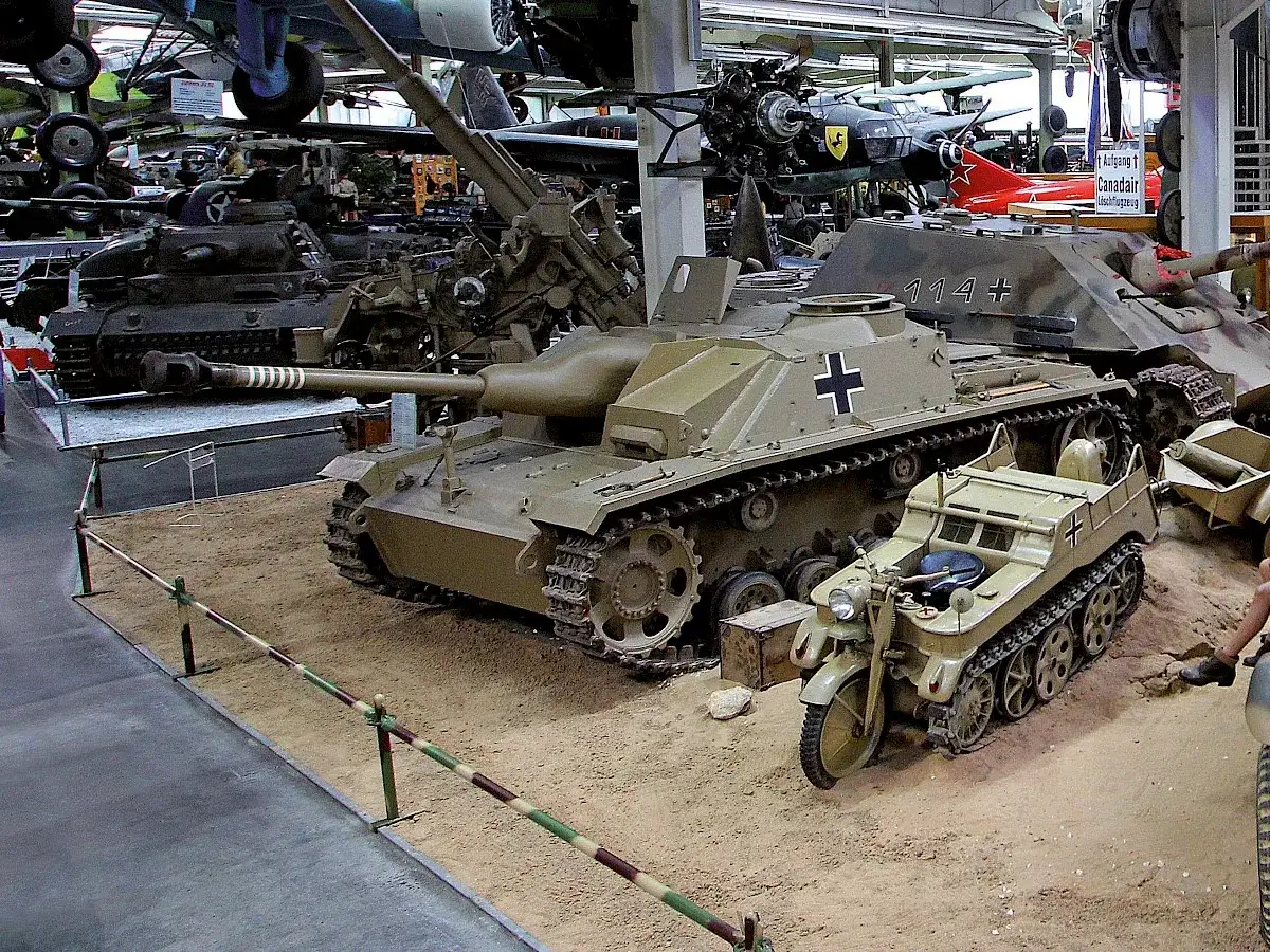Eine Sturmhaubitze 42 mit einem Kettenkrad davor, ausgestellt in einem Museum.